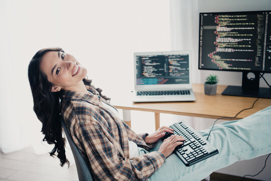 Smiling woman programmer working from home in a casual home office with laptop and monitor displaying code, typing on a keyboard with a relaxed posture