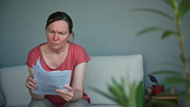 Anxious middle-aged woman reviewing debt notice and paperwork while sitting. Framed by plant foliage, conveying financial problems, budgeting challenges, household expenses. 4K
