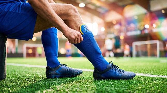 Football player adjusting shin guard before game. Man sitting on bench preparing to play soccer match on professional field. Athletic gear and equipment for sport competition activity