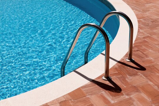 Gleaming metal pool ladder curves into brilliant blue water on brick patio