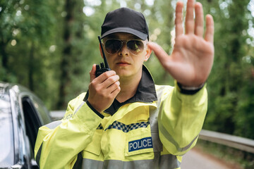 Male police officer in yellow reflective jacket and cap holds a radio while signaling to stop on a forest road with a patrol car in the background © standret