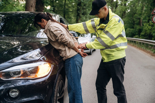 Law enforcement officer in reflective jacket arrests woman in casual clothing beside a black car on a tree-lined road during daylight