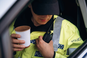 Police officer in yellow uniform holding a coffee cup while using a radio inside a patrol car, wearing a cap and visible police insignia on the jacket © standret