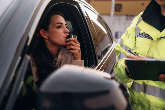 Woman in a car using a breathalyzer device while a police officer takes notes, urban setting with blurred background elements visible