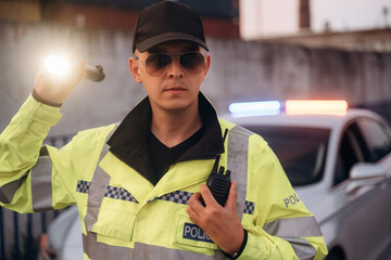 Male police officer in reflective yellow jacket holding flashlight and radio, standing near patrol car with flashing lights in urban environment during evening © standret