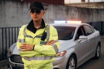 Male police officer in reflective yellow uniform stands confidently with arms crossed beside a silver patrol car with flashing lights in an urban setting © standret