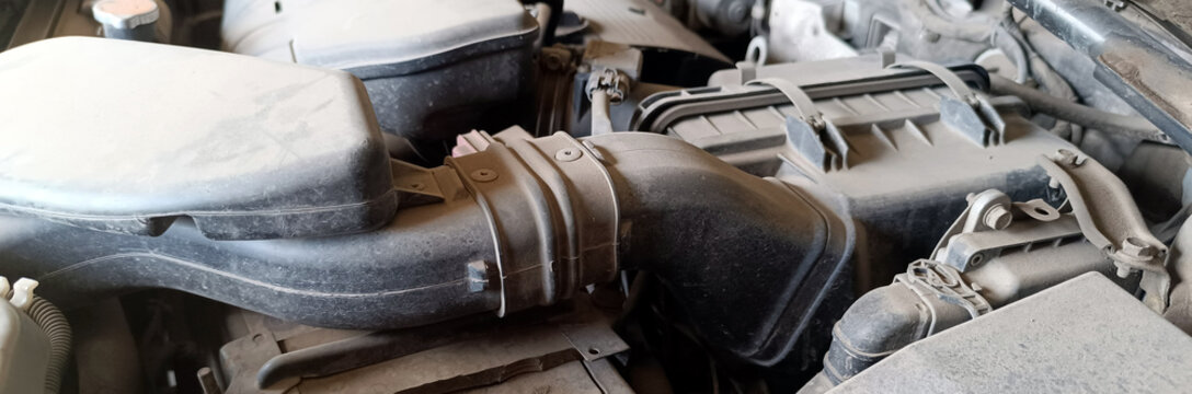 Detailed close-up shot of a dirty, dusty car engine bay, featuring a large black plastic air intake hose connected to a filter box. Various mechanical and electrical parts are visible