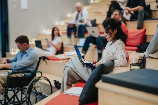 A group of diverse students study and take notes in a tiered lecture space. Several learners use laptops and notebooks while one student in a wheelchair participates