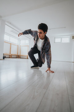 A young male dancer practices a low break dance move on a clean wooden studio floor. He balances on one hand in a spacious, light-filled rehearsal room