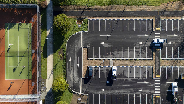 Aerial view of people playing tennis on a green court next to a modern parking lot with white directional arrows and parked cars in Nelson, Nelson Region, New Zealand.