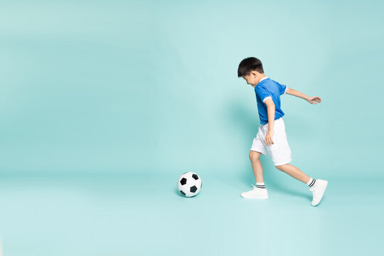 Full-length side view of a happy Asian boy in a blue jersey kicking a soccer ball, practicing sports on a mint green studio background with copy space. Active lifestyle and childhood football concept