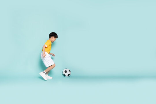 Side view of a cheerful Asian boy in a yellow jersey playing with a soccer ball, practicing football on a mint green studio background with copy space. Active lifestyle and childhood sports