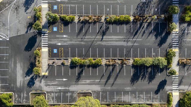 Aerial view of an empty parking lot with blue disabled parking spaces and green trees casting long shadows on the asphalt in Nelson, Nelson Region, New Zealand.