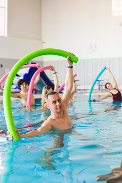 Group of people stretching with colorful noodles during an aqua aerobics class in an indoor swimming pool