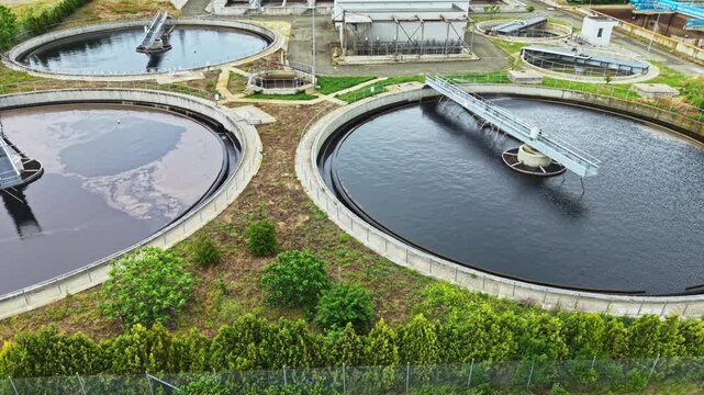 Aerial view wastewater treatment plant showing circular clarifiers and rotating scrapers, dark sludge basins bordered by green vegetation, concrete walkways,
