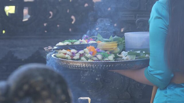 Slow motion: Balinese Hindu offering ceremony, with a woman placing flowers and incense on a ceremonial tray. Rising smoke creates a spiritual atmosphere during a temple ritual in Bali, Indonesia