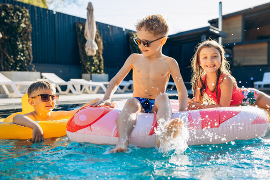 Three smiling children wearing sunglasses and swimsuits playing with inflatable floats in a sunny outdoor swimming pool, splashing water