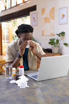 African man sneezing into tissue while working on laptop at home dining table with medicine