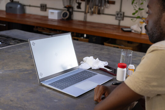 African American man sitting at island using blank laptop, pills, used tissues, copy space