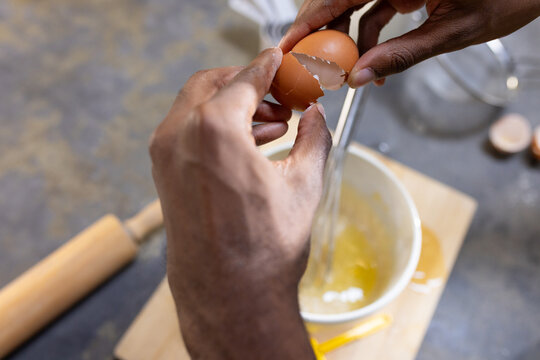 African adult male cracking brown egg into ceramic mixing bowl with metal whisk on cutting board