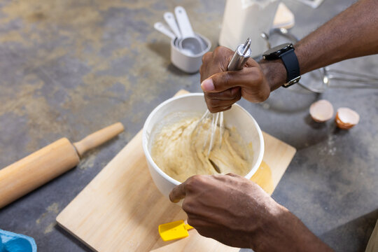 Man whisking batter with metal whisk in mixing bowl on kitchen counter, holding yellow spatula