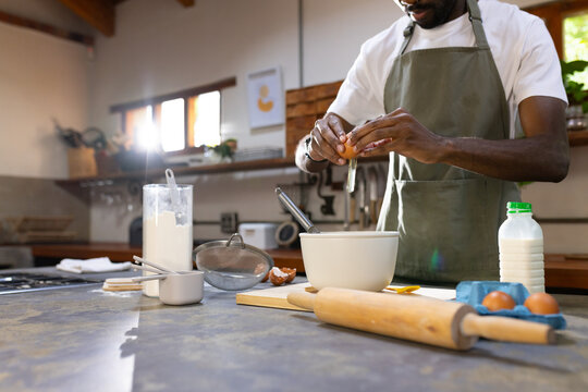 Adult African man cracking egg into mixing bowl at kitchen counter wearing green apron, copy space