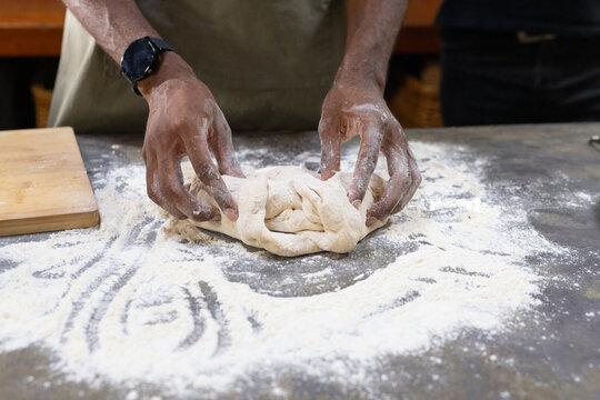 African American man shaping ball of dough on floured bench in bakery kitchen wearing apron