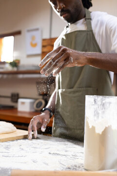African man wearing olive-green apron sprinkling flour at kitchen counter with dough and flour jar