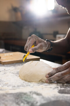 Adult African American man scoring round loaf with yellow-handled lame on floured wooden board