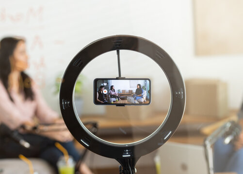Diverse female colleagues recording discussion in lounge with smartphone mounted in LED ring light