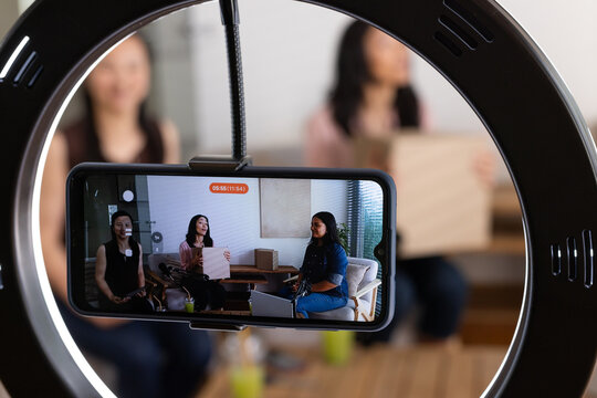 Three female hosts sitting on sofas at home, recording with ring light and smartphone showing board