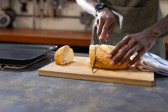 African man slicing crusty loaf with serrated knife on stone countertop, wearing green apron