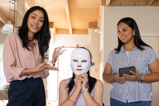 Diverse female friends presenting skincare demo at home with white LED mask, tablet, pink blouse