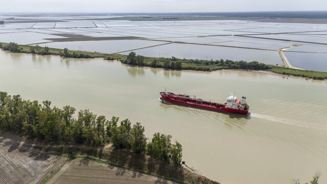 Aerial view of a red cargo ship navigating the Guadalquivir river past flooded rice fields and green riverbanks in Andalusia, Spain.