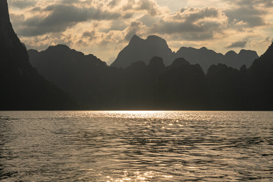 Golden sun glints dance on rippling water at sunset, silhouetting dramatic karst mountains against a cloudy sky in a serene natural landscape.