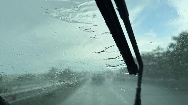 Rain pours down heavily on a busy highway. The windshield wipers move back and forth to clear the water. Cars drive in the rain with limited visibility in a city setting.