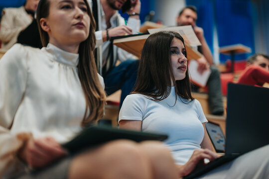 University students listens attentively during a lecture, with one student taking notes on a laptop. The classroom scene shows focused young adults studying and engaging in a campus seminar