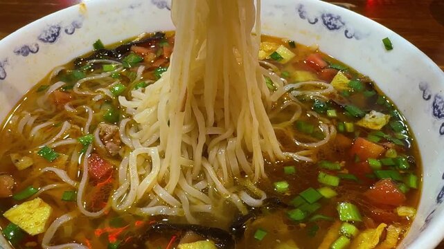 Person using chopstick to lift noodle from hot soup in bowl close up. Asian dish closeup with broth, vegetable and meat served for lunch. Traditional cuisine, healthy meal and dinner food culture.
