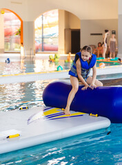 Smiling young girl wearing a life vest sitting on top of an inflatable bridge section of a water obstacle course in a public pool
