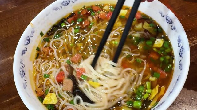 Person using chopstick to lift noodle from hot soup in bowl close up. Asian dish closeup with broth, vegetable and meat served for lunch. Traditional cuisine, healthy meal and dinner food culture.