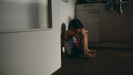 Lonely child sitting on kitchen floor with head down in corner expressing sadness isolation fear and emotional distress at home environment conveying vulnerability and neglect © Marco