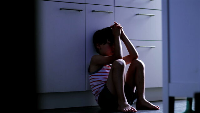 Child sitting on kitchen floor with hands clasped over head expressing overwhelming stress anxiety and childhood trauma in domestic environment suggesting fear and emotional breakdown