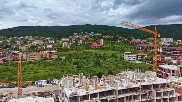 Tower crane above suburban green hillside under overcast sky, multi story concrete frames rising beside rows of new houses, busy worksite energy signaling