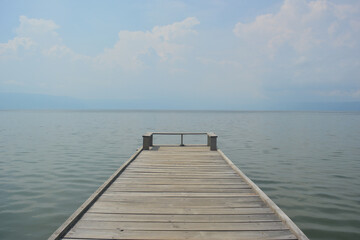 Obraz premium Wooden pier stretches from the bottom center into a calm body of water that meets the horizon under a soft, hazy sky emphasizes serenity and reflection for peaceful transitions, contemplative solitude