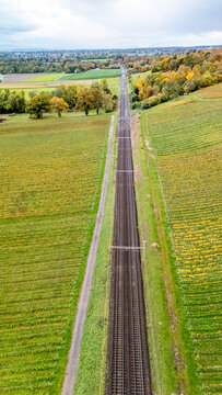 Aerial view of straight railway tracks stretching through lush vineyards and autumn-colored trees under a cloudy sky in Crans VD, Vaud, Switzerland.