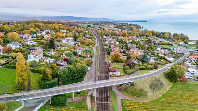 Aerial view of a railway line crossing through a residential neighborhood with autumn foliage and a road bridge near Lake Geneva Crans VD, Vaud, Switzerland.