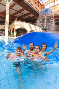 Happy family portrait smiling and waving hands in an indoor swimming pool with water mushroom fountain at a water park