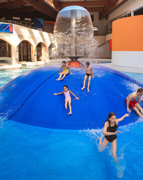 Large group of happy people jumping from a blue water mushroom fountain dome at an indoor water park