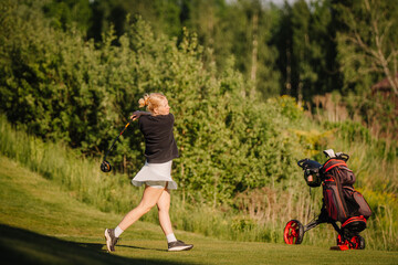 Fototapeta premium Young female golfer swings club on green course with golf bag nearby, surrounded by lush trees and grass under bright sunlight in a recreational setting
