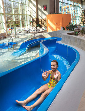 Happy young girl waving hands while sliding down a blue water slide at an indoor water park